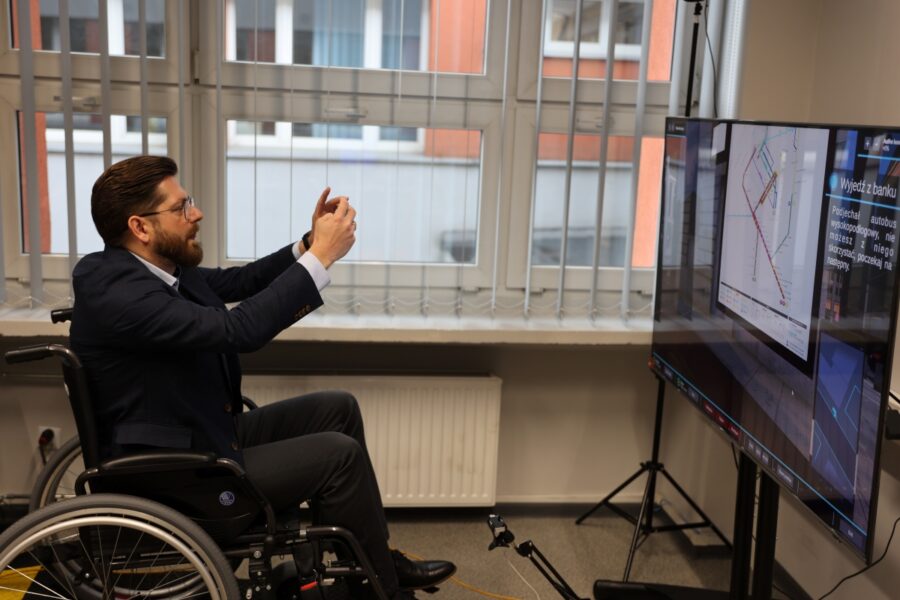 Zdjęcie przedstawia mężczyznę na wózku inwalidzkim przed nim jest telewizor na telewizorze jest włączony program szkolenia, mężczyzna robi zdjęcie programu w celu dokumentacji