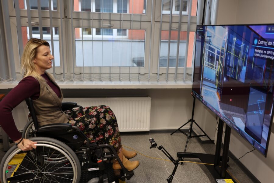 Zdjęcie przedstawia kobietę na wózku inwalidzkim przed nią jest telewizor na telewizorze jest włączony program szkolenia,