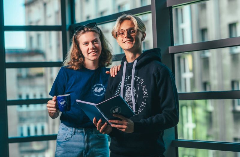 dwóch studentów stoi i patrzy się w obiektyw. Jeden trzyma w ręku notatnik, a studentka trzyma kubek. 