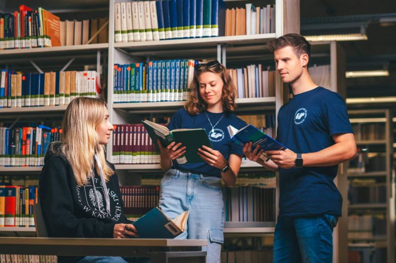 jedna studentka siedzi przy stoliku z książką w ręku i patrzy na dwie osoby które stoją, one również mają książki w dłoniach