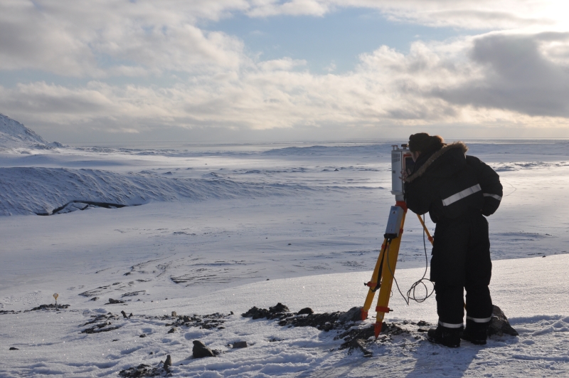 Kobieta ubrana w zimowy kombinezon patrzy przez aparaturę badawczą na odległe tereny arktyczne/Woman dressed in winter clothes is looking through research apparatus at distant Arctic areas