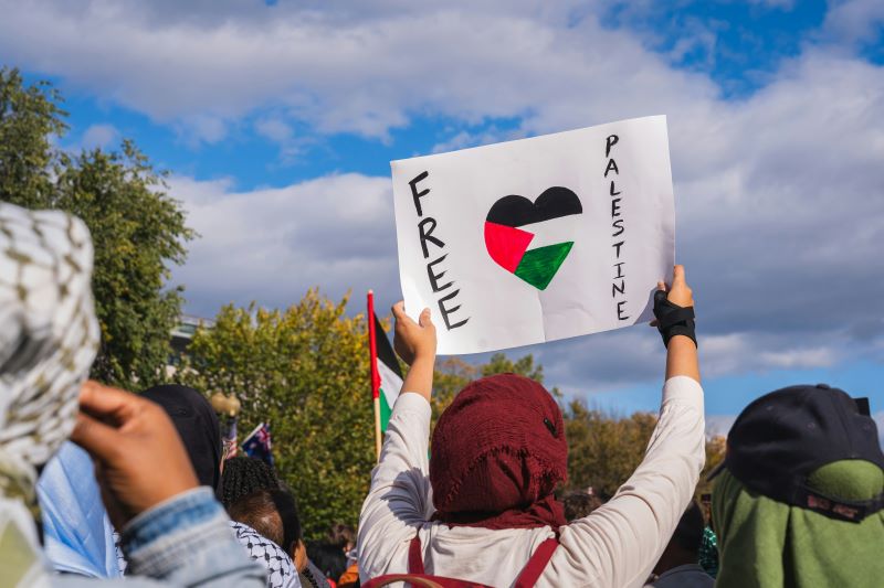 Tłum ludzi. Jedna osoba odwrócona tyłem trzyma w dłoniach kartę z napisem Free Palestine. Flaga Palestyny w kształcie serca