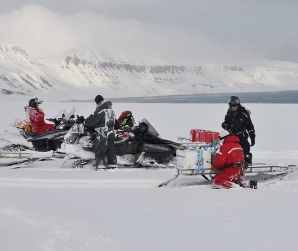Kilkoro naukowców na Spitsbergenie ubranych w czerwone i czarne kombinezony, na skuterach śnieżnych