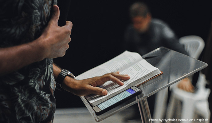 Osoba wygłaszająca wykład, zbliżenie na mównicę i otwartą książkę. Fot.  Nycholas Benaia on Unsplash //Person giving a lecture, close-up of the platform and open book