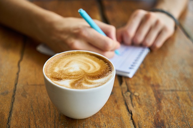 Na pierwszym planie kawa, na drugim ręka człowieka zapisującego coś długopisem w notesie