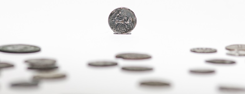 Kilkanaście rozrzuconych monet rzymskich na białym blacie, jedna z nich w tle w pozycji pionowej.