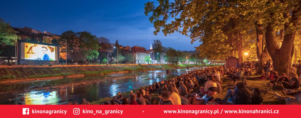 Uczestnicy przeglądu filmowego oglądają projekcję filmu, wydarzenie odbywa się w plenarze, ekan umieszczony jest po jednej stronie rzeki, uczestnicy siedzą na drugim brzegu  