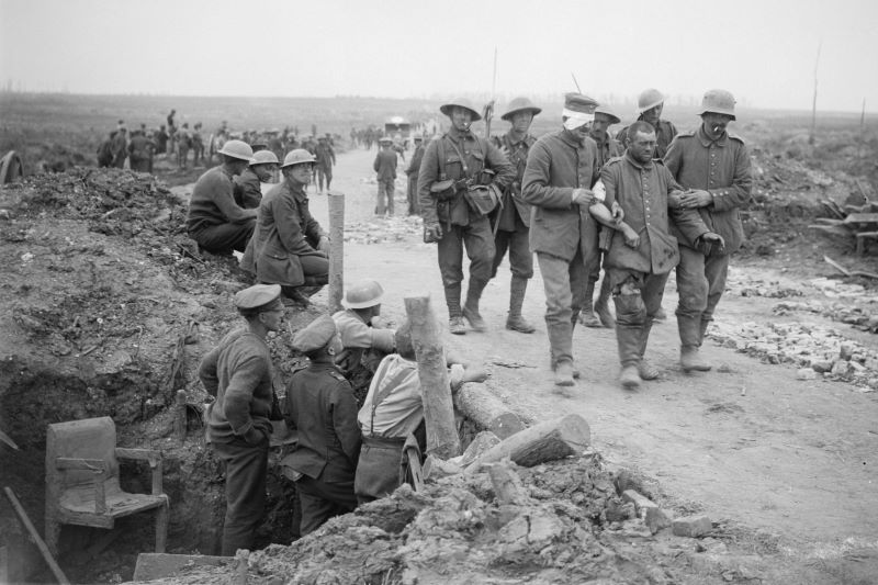 Zdjęcie z I wojny światowej ukazujące rannego żołnierza podtrzymywanego przez kolegów. Część osób idzie ścieżką za nimi, część siedzi wzdłuż drogi