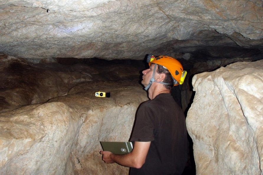 Dr Andrzej Tyc w jaskini, z kaskiem na głowie i laptopem w rękach przygląda się skałom