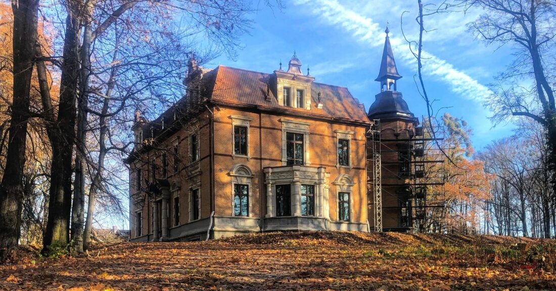 Pałac w Rzuchowie podczas jesieni. Wokół budynku leżą liście ze stojących obok drzew. Niebo z chmurami