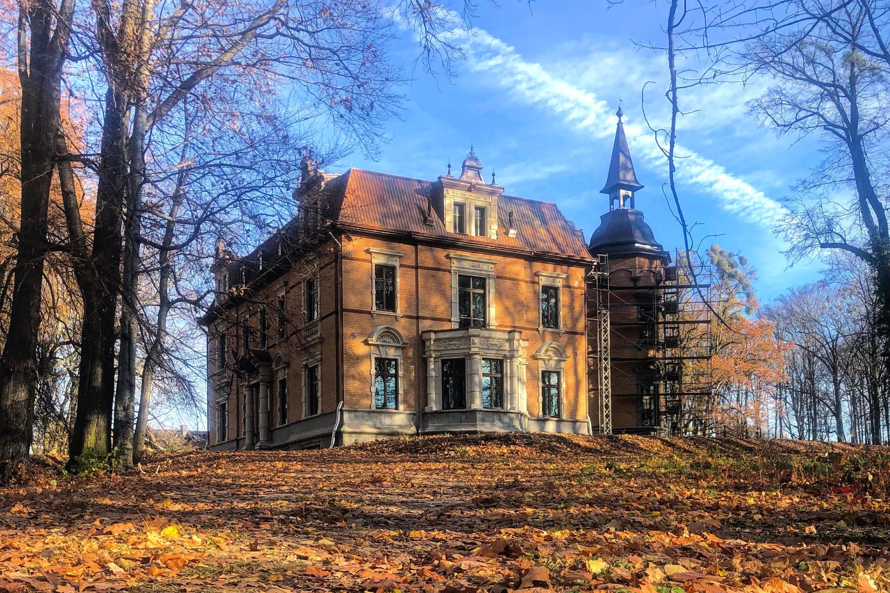 Pałac w Rzuchowie podczas jesieni. Wokół budynku leżą liście ze stojących obok drzew. Niebo z chmurami