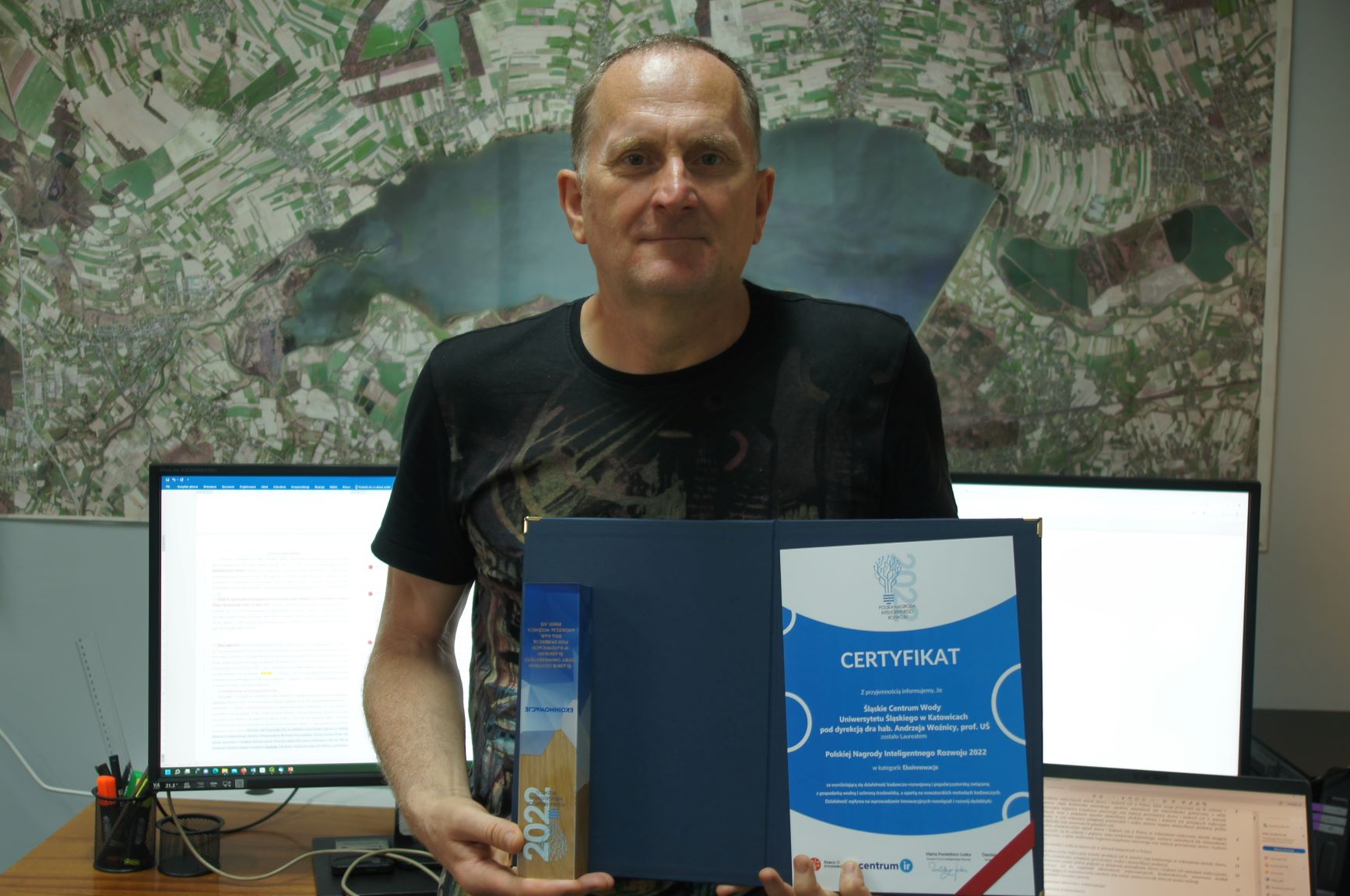 Prof. Andrzej Woźnica prezentuje nagrodę i dyplom. Za nim mapa na ścianie i świecące się monitory komputera