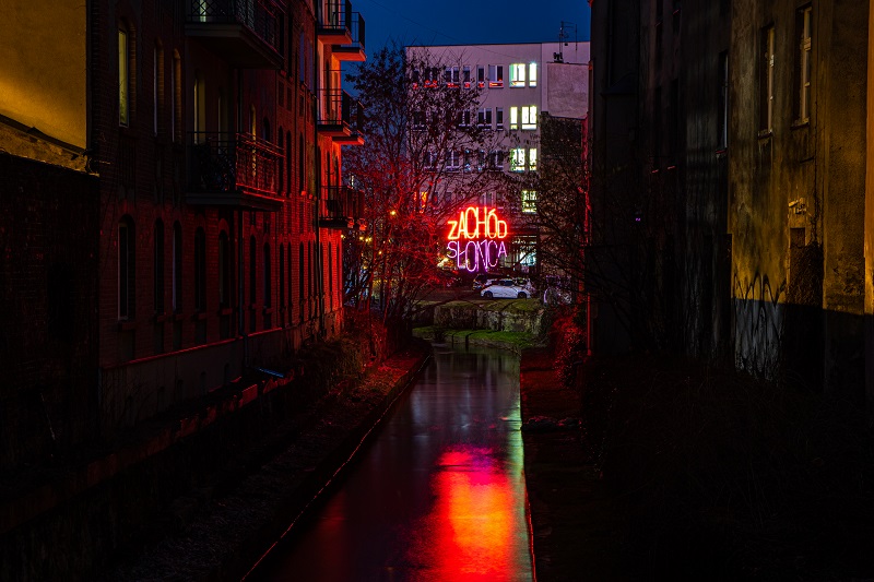 Rawa przepływająca pomiędzy budynkami. Na środku świecący neon z napisem Zachód Słońca
