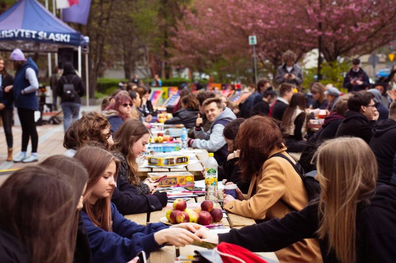 grupa studentów siedząca przy jednym długim stole na zewnątrz