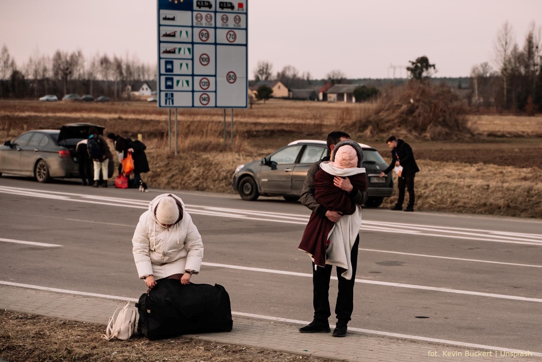 Ludzie stojący na granicy z torbami. Na pierwszym planie kobieta pochyla się nad torbą, po prawej stronie stoi mężczyzna z dzieckiem na rękach. Za nimi widać dwa samochody i ludzi wokół nich, znak drogowy oraz pola