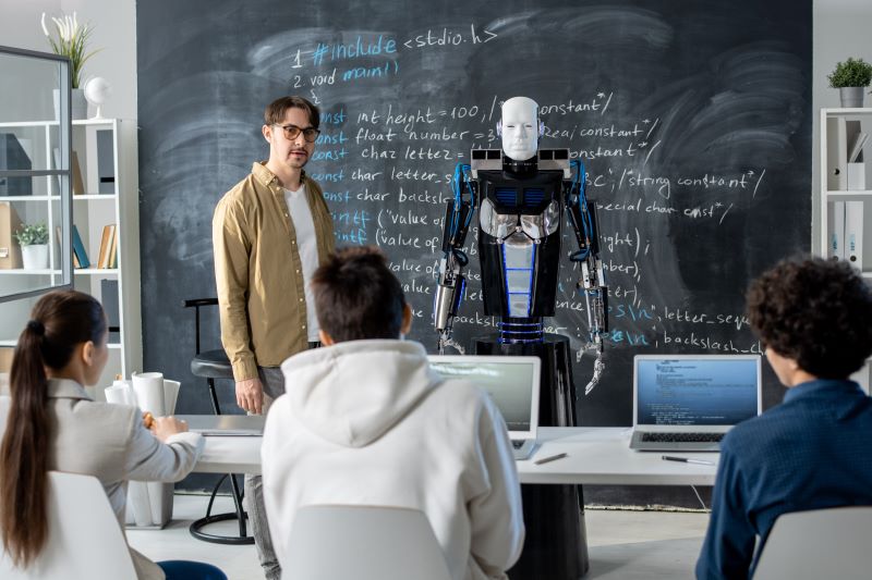 Nauczyciel stoi przy zapisanej tablicy, obok niego robot. Dzieci wpatrują się w nich/Teacher standing by the blackboard with a human-like robot next to him. Children are looking at them