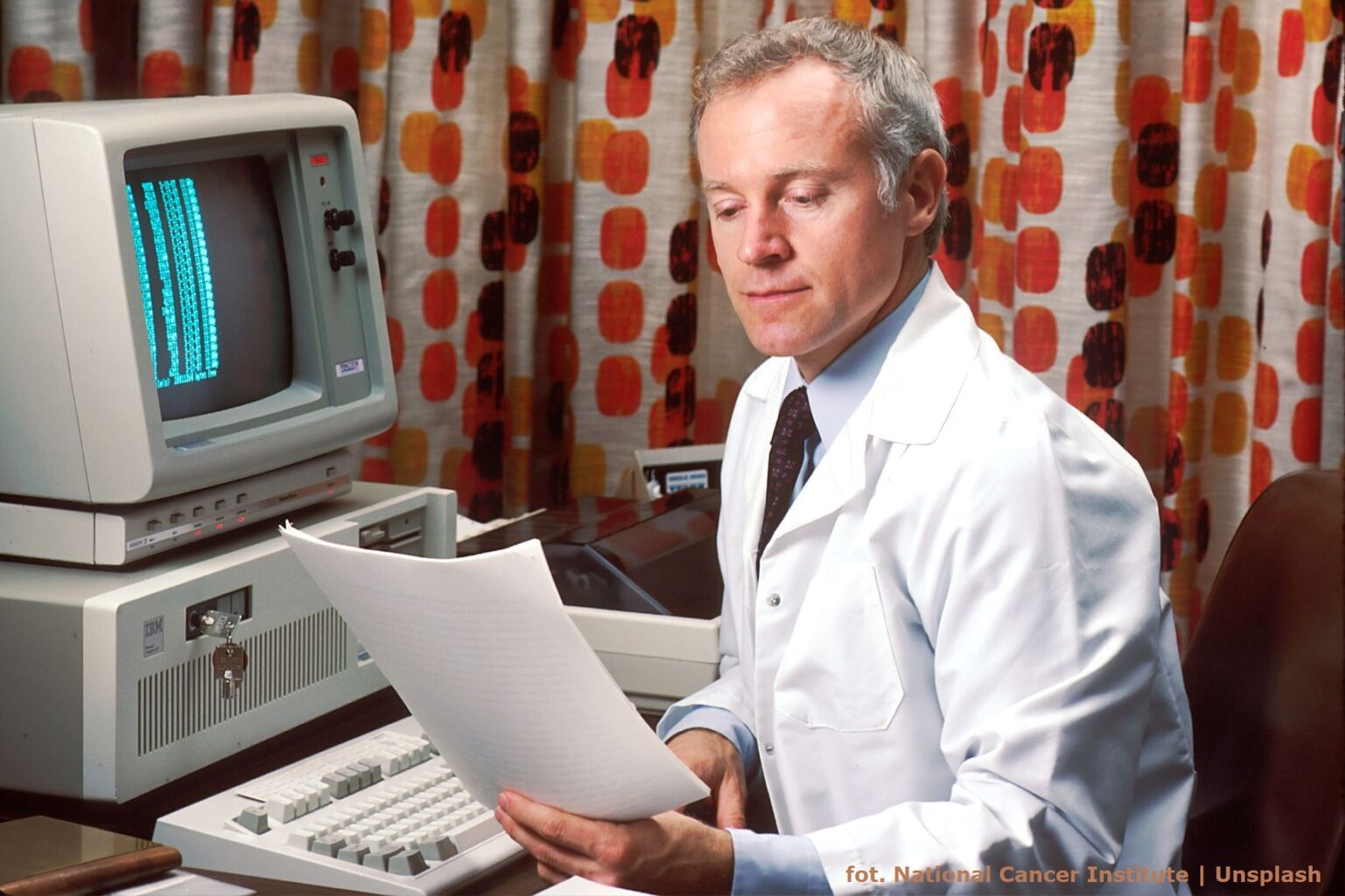 Naukowiec w fartuchu siedzi przy biurku trzymając kartki papieru. Przed nim monitor komputera w starym stylu