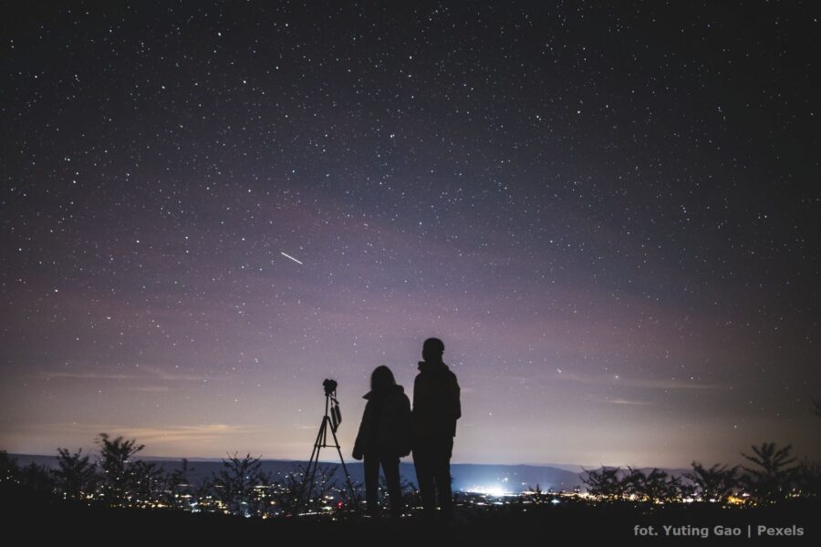Para z aparatem na statywie stoi i obserwuje nocne niebo. W tle rozświetlone miasto. Główny widok na rozgwieżdżone niebo