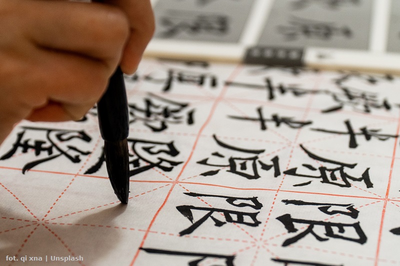 Dłoń trzymająca pędzel do kaligrafii. Na papierze widać napisane chińskie znaki