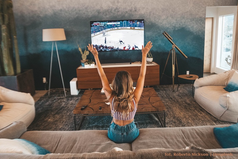 Dziewczyna siedząca przed telewizorem dziewczyna z uniesionymi rękami do góry. W telewizorze widać rozgrywany mecz hokejowy