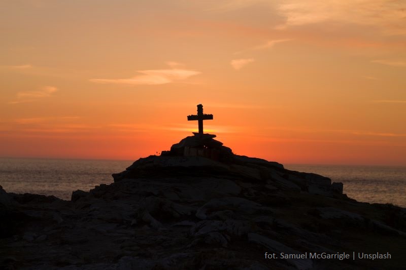 Zachód słońca nad morzem, na pierwszym planie wzgórek, na którym stoi krzyż