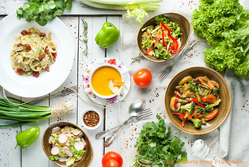 Widok z góry na różne wegańskie potrawy w talerzach, półmiskach i pojedyncze warzywa oraz owoce wprost na stole