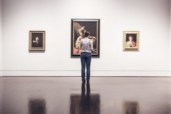kobieta stojąca tyłem ogląda wystawę, na ścianie wiszą trzy obrazy