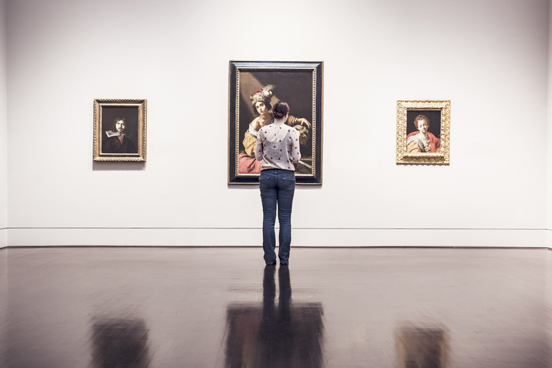 kobieta stojąca tyłem ogląda wystawę, na ścianie wiszą trzy obrazy