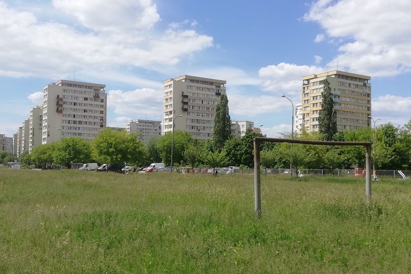 Osiedlowe boisko z jedną bramką, boisko porośnięte trawą, w oddali widać bloki mieszkalne
