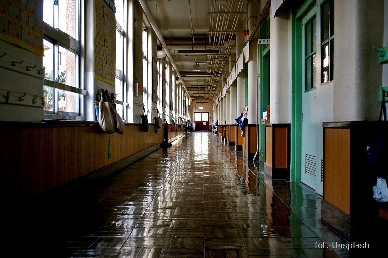 Widok pustego korytarza szkolnego. Po prawej stronie drzwi do sal lekcyjnych, po lewej okna oświetlające pomieszczenie./An empty school corridor with classrooms from the right and windows from the left.