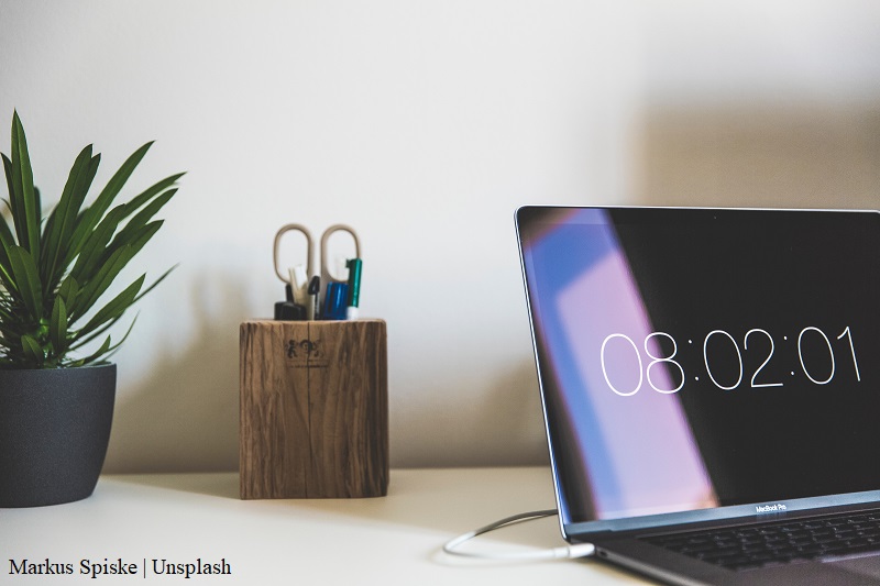 na biurku leżą: laptop z wyświetloną godziną, roślina w doniczce, przybornik z długopisami i nożyczkami / laptop, plant and office supplies on the table