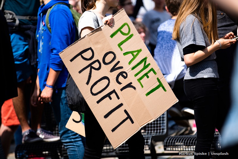 Widok na trzymany w ręce karton z hasłem „Planet over profit” trzymany przez dziewczynę w tłumie