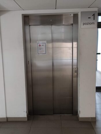 Metalowe drzwi windy, widok z zewnątrz. po prawej panel przywołania windy. W prawym górnym rogu metalowa tabliczka z napisem -1 poziom.