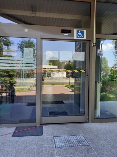 Przeszklone drzwi do budynku oznakowane kontrastami. Przed drzwiami wycieraczka. Nad drzwiami piktogram osoby na wózku inwalidzkim.