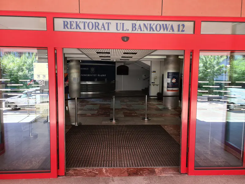 Rama drzwi czerwona. Drzwi szklane, transparentne, otwarte. Nad drzwiami napis: „Rektorat ul. Bankowa 12”