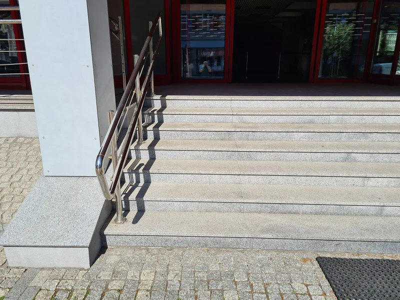 Szare schody przed wejściem. Przed schodami podłoże z kostki brukowej i wycieraczka. Po lewej stronie schodów poręcz. Za poręczą kolumna/Grey stairs in front of the entrance. Cobblestone surface in front of the stairs and a doormat On the left of the stairs is the platform for wheelchairs. Column behind the handrail.