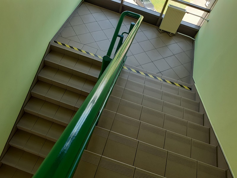 Widok od góry na klatkę schodową. Beżowe schody zakończone taśmą kontrastową, zielona poręcz schodowa/View from the top of the staircase. Beige stairs finished with a contrasting tape, a green handrail.
