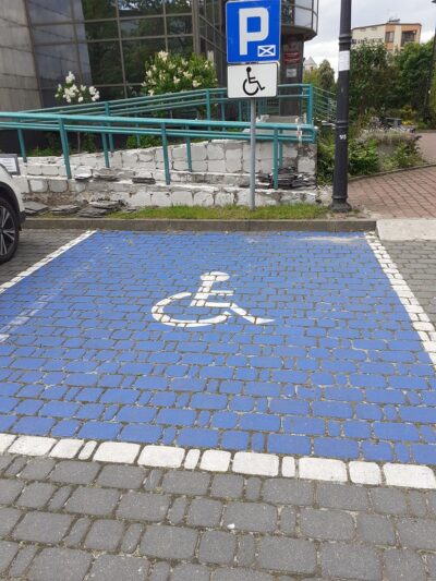 Miejsce parkingowe dla osób niepełnosprawnych – niebieska nawierzchnia z białym znaczkiem osoby na wózki inwalidzkim. Przed nim znak drogowy P i oznaczenie wózka inwalidzkiego. W tle widać podjazd i budynek.