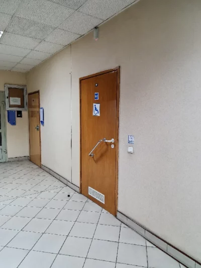 Korytarz po prawej stronie widać drewniane drzwi. Na drzwiach niebieskie oznaczenie WC i piktogram osoby na wózków. Pod klamką drzwiową dodatkowy uchwyt. Po prawej stronie drzwi, na wysokości klamki włącznik z napisem „zgaś światło”