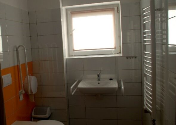 Widok wnętrza toalety. Z lewej ustęp otoczony uchwytem, pojemnik na papier, pod nim kosz. Na wprost okno, pod nim umywalka z pochwytami po oby stronach. Po prawej grzejnik.