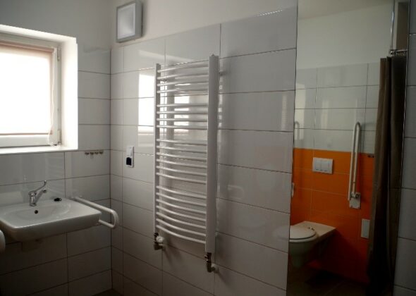 Widok wnętrza łazienki. Po lewej umywalka, okno. Po prawej grzejnik łazienkowy. Dalej powieszone jest lutro, w którym odbija się miska ustępowa i kawałek kotary prysznicowej.