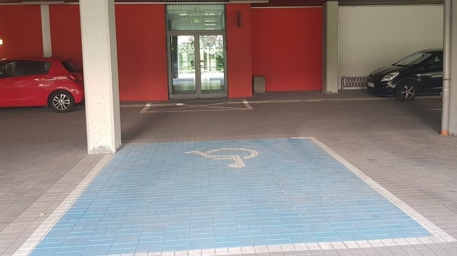 Parking podziemny. Na pierwszym planie widać oznakowane miejsce parkingowe dla osób niepełnosprawnych. W oddali wejście do budynku