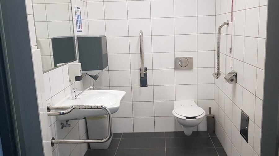 Wnętrze toalety. Po lewe drzwi wejściowe, umywalka z poręczą, przedłużoną baterią umywalkową oraz lustrem. Dalej podajnik na ręczniki, pod nim kosz. Na wprost miska ustępowa i dwa uchwyty./Interior of the toilet. On the left the entrance door, washbasin with a handrail, extended washbasin tap and mirror. Next is the towel feed, with a bin under it. Opposite is the toilet bowl and two holders.