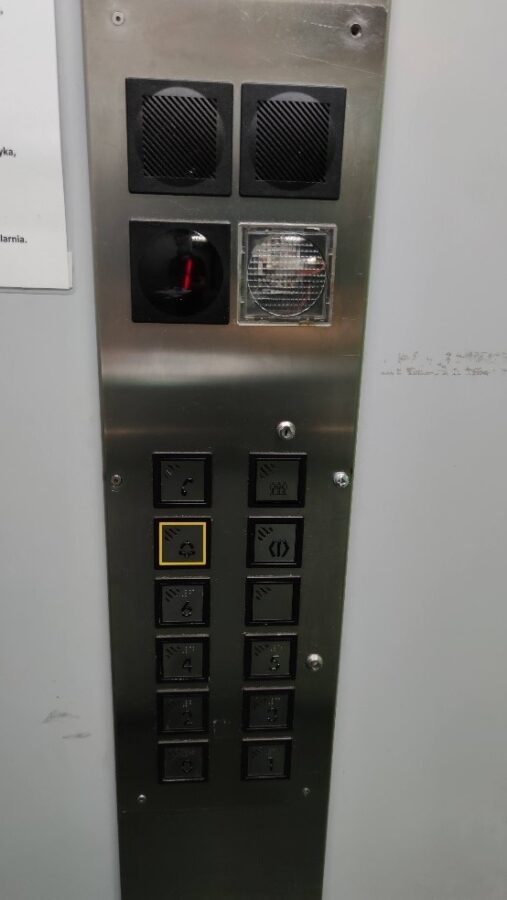 Panel sterujący windą. Na górze cztery większe kwadraty. Dwa górne to głośniki, na dolnym lewym kwadracie znajduje się świetlny wyświetlacz z numerem piętra, prawy dolny kwadrat to lampka. Poniżej są dwa rzędy kwadratowych mniejszych klawiszy z wypukłymi oznaczeniami. Lewy górny klawisz to słuchawka telefonu, poniżesz dzwonek, górny prawy przycisk pokazuje grupę ludzi, drugi od góry z prawej to przycisz znaku symbolizujący otwieranie drzwi. Pozostałe przyciski to numery w wypukłym czarnodudku i alfabecie Braile’a./Lift control panel Four larger squares at the top. The two squares at the top are loudspeakers, the bottom left square contains a light display with the floor number, the right bottom square is a lamp. Below are two rows of smaller keys with convex labels. The left upper key is a telephone receiver, below is the call-bell, the upper right button shows a group of people, the second button from the top on the right is the button symbolising door opening. The other buttons are numbers in convex common scimitarbill and Braille alphabet.