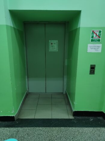 Drzwi zewnętrzne do windy umieszczone we wnęce. Panel przywoławczy windę umieszczony przed wnęką. Nad panelem są dwie tabliczki. Ściana na dole ma kolor ciemno zielony, wyżej jasnozielony.