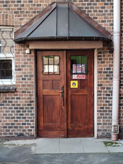 Brązowe drewniane drzwi, wejście bez progu, podłoże z kafelek i asfaltu.