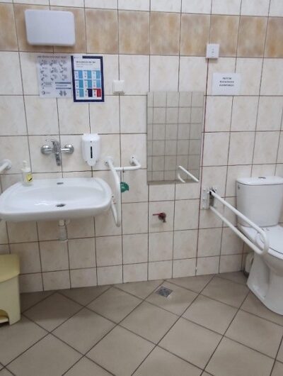 Wnętrze toalety dla osób niepełnosprawnych. Po lewej umywalka po prawej toaleta. Na ścianie z lewej strony, podajnik na [ręczniki papierowe. Po prawej stronie na ścianie podajnik na papier toaletowy.