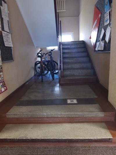 Wnętrze budynku. Dwa schodki prowadzące do klatki schodowej. Po prawej schody prowadzące na górę Na ścianie tablica informacyjna. Po lewej pod obniżeniem schodowym stoją dwa rowery.