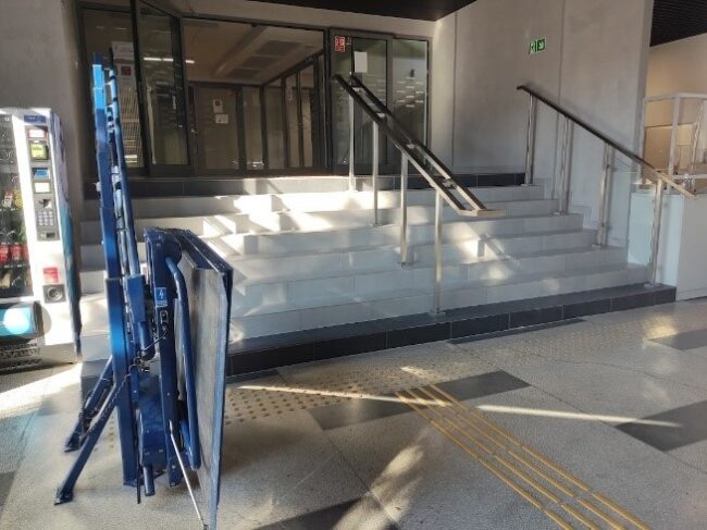 Wnętrze budynku. Przed schodami na wyższy poziom budynku znajduje się ścieżka naprowadzająca i pole uwagi. Po lewej stronie schodów platforma dla wózka inwalidzkiego. W środku i po brzegach schodów metalowe poręcze. Za schodami przeszklone automatycznie otwierane drzwi.