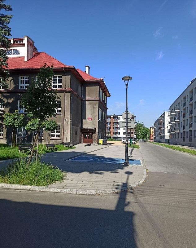 Skrzyżowanie ulic Będzińskiej i Staropogońskiej oraz budynek Wydziału Nauk Ścisłych i Technicznych – szary stary budynek z dużymi białymi oknami i czerwonym spadzistym dachem. W okolicy widać inne budynki.